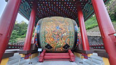 The image shows a large, elaborately decorated drum housed within a traditional Korean pavilion in Godang-ri, South Korea. The pavilion features vibrant red pillars and a patterned ceiling with circular motifs. The drum itself is the central focus, adorned with intricate paintings of dragons and floral designs in a rich palette of colors. It rests on a wooden platform, supported by red wooden braces. Steps lead up to the pavilion, and a blue rope barrier is visible, indicating a public viewing area. The background consists of a green hillside with scattered trees and a stone wall. The scene is brightly lit, suggesting it was taken during the daytime. There are no people visible in the image.