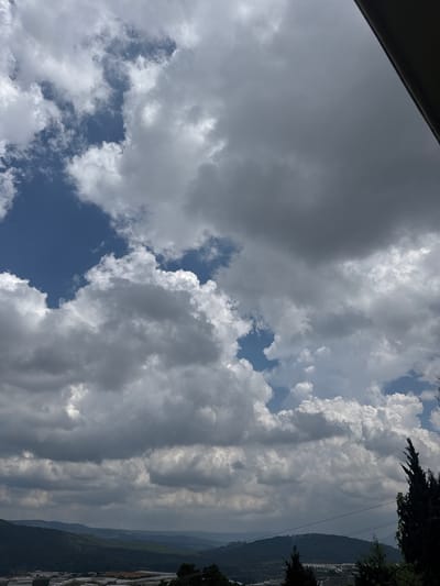 The image is a vertical shot of a partly cloudy sky over a landscape in Đa Lộc, Vietnam. The sky is filled with a mix of fluffy white cumulus clouds and darker, more diffused grey clouds. Patches of bright blue sky are visible between the clouds. In the foreground, the top edge of a dark structure, possibly a roofline or overhang, is visible in the upper right corner. The middle ground shows rolling hills covered in dark green vegetation. In the lower portion of the image, a cluster of greenhouses with white roofs and frames can be seen, indicating agricultural activity. A few dark green evergreen trees are visible along the right edge and scattered throughout the landscape. The overall impression is of a bright, overcast day with a hint of agricultural development in a hilly region.