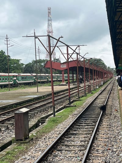 This is a photograph taken at the Sylhet Railway Station in Sylhet, Bangladesh. The image shows a railway track in the foreground, with a black dog walking on the tracks. To the left, a train with green and white carriages is parked at the station. Further left, trees and a telecommunication tower are visible. To the right, a covered platform area with red pillars and a red roof extends along the track. The sky is cloudy.