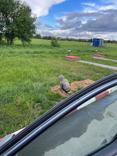 The image shows a grassy field in Бавыкино, Russia. The field contains two red and white barrier markers. In the middle ground, there is a small blue and white portable toilet. In the foreground, a young child wearing a grey hooded sweatshirt with lettering is kneeling near a pile of dirt. The scene is viewed through the partially open window of a vehicle. A red and white curb is visible on the right side of the image. The sky is partly cloudy.
