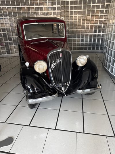 A vintage, dark red and black Fiat 500 Topolino is parked indoors on a white tiled floor. The car faces forward, with its chrome grille prominently displayed. A license plate reading "18 CE" is visible on the front. The background consists of a wall made of translucent glass blocks, which are illuminated from behind. To the right, a curved wall of similar glass blocks is also present. The lighting appears to be artificial, casting soft shadows on the car and floor. The overall scene suggests a display or storage environment for classic automobiles. The car is a Fiat 500 Topolino, manufactured in Italy, and the city of Calvi Risorta in Italy is mentioned as the location, implying this might be a car show or private collection in that area.