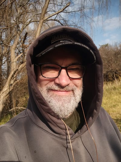 This is a close-up, rotated selfie of an older white male with a grey beard and glasses. He is wearing a dark grey hooded sweatshirt, with the hood pulled up. The background is blurred, showing dry trees and a bright blue sky, suggesting an outdoor setting during the day. The man appears to be looking directly at the camera, with a neutral expression. The image has a warm, natural light, indicating it was likely taken on a sunny day. Text is visible on the inside rim of the hood: "COMPLY. TOUGH. LONGO."