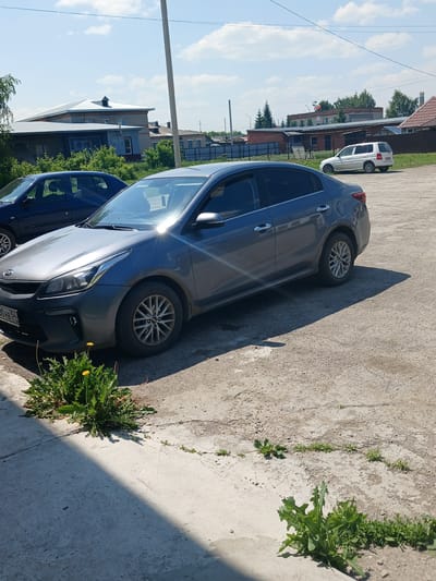 The image depicts a gray Kia Rio sedan parked on a gravel and concrete lot. The car is positioned in the foreground, with its front end angled towards the left of the frame and its rear towards the right. A dark blue sedan is partially visible to the left of the Kia. In the background, there are several buildings, including a light-colored structure with a metal roof and a longer brick building. Trees and foliage are scattered throughout the scene, and the sky is visible with scattered clouds. The ground is uneven, composed of gravel and cracked concrete, with patches of green vegetation, including dandelions, growing in the cracks. A single utility pole stands in the mid-ground. The scene appears to be a daytime outdoor setting, likely in a residential or industrial area of Prokudskoye, Russia, given the presence of typical Russian architecture and vehicles.