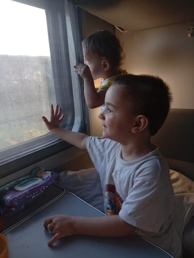 Two young children are looking out of a train window in Zagarancea, Moldova. The older child, a boy, is in the foreground, wearing a white t-shirt with a cartoon character on it. He is looking to the right, with his mouth slightly open. His left hand is resting on a tray table in front of him. Behind him and slightly to the left, a younger child, a girl, is looking out the window to the left, with her hand near the window screen. She is wearing a yellow and white top. The train compartment appears to be a sleeping berth, with bedding visible behind the children. A package of "Baby Wet Wipes" is on the tray table. The lighting suggests it is either dawn or dusk, with a bright sky visible through the window.