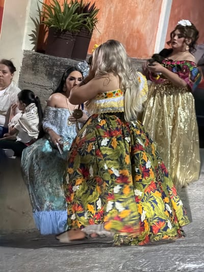 Four women pose in traditional Mexican dresses. The dresses feature a variety of colors and floral patterns with varying textures. One woman wears a shiny, golden dress. A woman with blonde hair leans over, adjusting the dress of another. Another group of two women is seated on a stone staircase and one is wearing a white shirt. A potted plant sits on the top step, near the women.