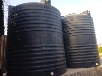 Two large, black, ribbed water tanks stand side-by-side on a concrete base. The tank on the left has a notable spiderweb with a few strands visible on its upper surface. Both tanks bear the word "MACLEAN" in raised lettering. The surrounding area includes parts of a corrugated metal structure on the left, greenery in the background, and a bright sky above. The tanks appear to be located outdoors.