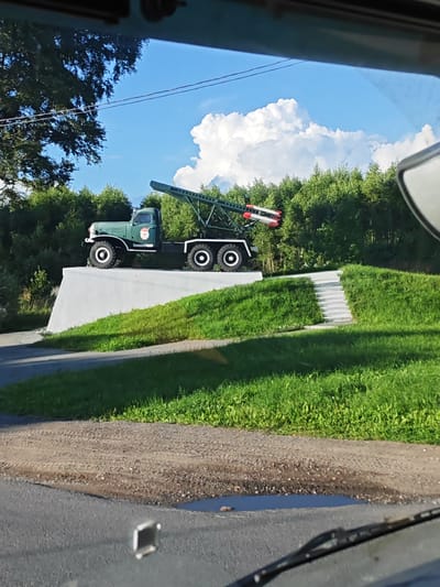 This is a forensic, matter-of-fact description of an outdoor scene. The image depicts a green military truck with a missile launcher mounted on its bed, positioned on top of a white concrete platform. The truck is facing left and has its tires turned slightly. Two red and white missiles are visible on the launcher. The platform is situated on a grassy hill, with a set of concrete stairs leading up to it from the right. The area around the platform is covered in green grass. In the background, a dense line of trees forms a backdrop, with a bright blue sky and white clouds above. The scene appears to be a monument or memorial. The image is taken from inside a vehicle, as suggested by the presence of a windshield wiper and a reflection in the windshield. The location is identified as the city of Shornovo, Russia.