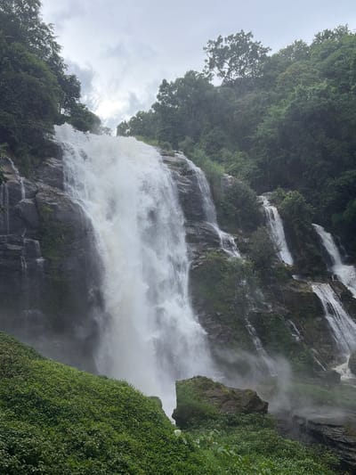 The image shows a majestic waterfall cascading down a rocky cliff face, surrounded by lush green vegetation and dense forest. The sky above is overcast with dramatic clouds, hinting at a potentially humid or rainy atmosphere. The water is white and frothy, creating a powerful visual of nature's force. The foreground is dominated by a steep, verdant slope covered in thick, low-lying plants. The waterfall itself appears to be multi-tiered, with water flowing down in various streams and rivulets over the dark, wet rock. Some mist is visible rising from the base of the falls, obscuring parts of the lower cliff. Trees with dense canopies line the top of the cliff and the surrounding hills, indicative of a tropical or subtropical environment. The scene is likely located in a natural park or protected area, characteristic of regions like Ban Sop Hat, Thailand, known for its natural beauty and waterfalls. No people are visible in the image.