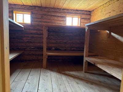 The image depicts the interior of a rustic wooden structure, likely a bunkhouse or sleeping quarters, with two small windows allowing natural light to enter. The walls are constructed from rough-hewn logs, giving the space a primitive and aged feel. The ceiling is also made of wooden planks, aligned horizontally. There are multiple levels of wooden bunks or shelves built into the structure, with visible supports and shelving. The floor is made of unfinished wooden planks laid side-by-side. The lighting suggests a daytime scene, with sunlight casting shadows across the floor and bunks. The overall impression is one of simple, functional accommodation, likely in a remote or historical setting. The provided context indicates this location is in Ust'-Pyzas, Russia.
