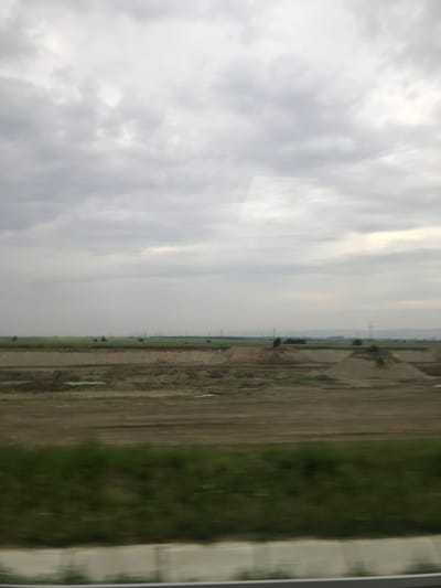 The image depicts a vast, partially developed rural landscape, likely viewed from a moving vehicle given the motion blur in the immediate foreground. The upper two-thirds of the frame is dominated by an overcast sky filled with uniform grey clouds, with a slightly brighter area towards the upper right suggesting diffused light.
Below the horizon, an expansive agricultural area unfolds, characteristic of the region around Adjud, Romania. This land is segmented into large patches of vibrant green fields, possibly cultivated crops or pastures, interspersed with broad sections of brown, freshly tilled earth. Across the green fields on the left, several dark, rounded objects resembling hay bales are scattered. Numerous utility poles with overhead lines traverse the distant landscape horizontally, indicating widespread electrical infrastructure. Faint, undulating hills or mountains are visible on the far horizon, particularly towards the right.
Closer to the viewer, in the midground, is a substantial area of disturbed earth exhibiting lighter brown and sandy hues. This area contains several large, conical mounds of earth or sand, indicative of excavation, construction activity, or stockpiled materials. Small, irregular puddles or bodies of water reflect the subdued light within this disturbed zone. The immediate foreground shows a blurred strip of dark green vegetation, likely grass, bordered by a light-colored, horizontally blurred linear structure, possibly a concrete road barrier or curb, further suggesting the viewpoint from a moving vehicle.
No human figures, animals, or specific actions are visible within the scene. The environment conveys a sense of quiet rural expansion or development under an overcast sky.