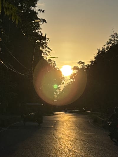 The image captures a sunset scene on a road in Ranong, Thailand. The sun, a bright, diffused orb, is positioned high in the sky, casting a warm, golden light that reflects off the wet asphalt road. Dense foliage lines both sides of the road, creating a natural canopy. A three-wheeled vehicle, commonly known as a tuk-tuk, is visible on the left side of the road, its form silhouetted against the bright background. The road appears to be a rural or semi-rural route, with power lines and streetlights visible along the sides. The overall atmosphere is serene and tranquil, characteristic of a tropical sunset.