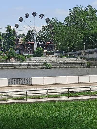 The image depicts a view of a Ferris wheel with dark gondolas against a bright sky with scattered clouds. The Ferris wheel is located in a park-like setting, with lush green trees in the background and a body of water in the foreground. Several buildings, some with signage including "VINCENZO", are visible behind the Ferris wheel. The area surrounding the Ferris wheel appears to be an amusement or recreational zone. The foreground shows a manicured lawn and a railing. The scene appears to be captured on a sunny day. The image is a photograph and does not contain any information about people or specific actions.