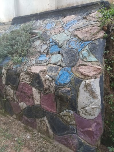 A close-up of a retaining wall constructed of large, irregular stones, painted in varying colors like blue, purple, pink, and gray. The wall appears weathered, with some moss or lichen growth. Vegetation is visible along the top edge and base of the structure. The construction has a rugged, hand-built appearance with thick mortar joints. The overall impression suggests an aged, possibly garden or roadside structure.