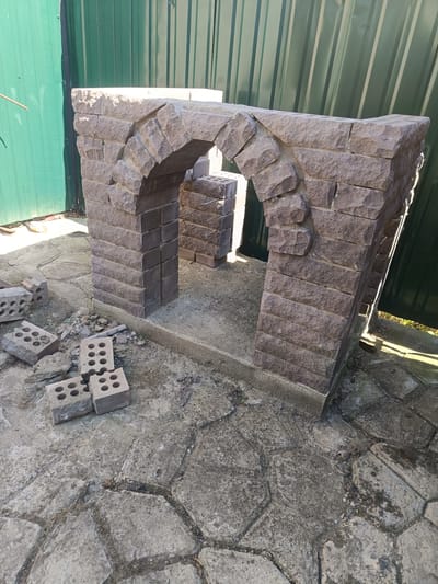 The image shows a partially constructed stone archway in Peremozhne, Ukraine. The archway is made of rough-hewn, reddish-brown stones. It stands on a concrete base, with loose bricks and debris scattered on the ground around it. Behind the archway is a green corrugated metal fence. The ground is paved with irregular concrete slabs. The scene suggests a construction or renovation project is underway.