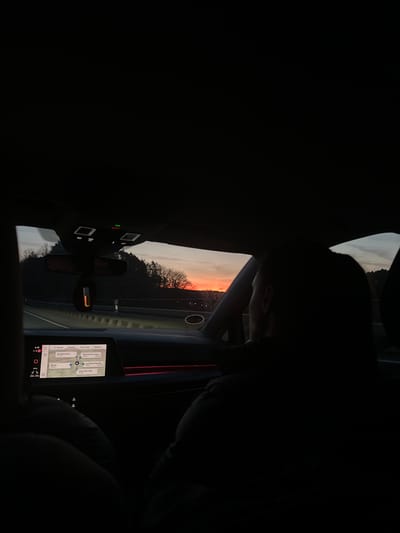 The image shows the interior of a vehicle, possibly a car, at dusk or dawn. The outside view through the windshield reveals a landscape with trees against a sky displaying colors of orange and gray, suggesting either sunrise or sunset. Inside the vehicle, a digital display screen is visible, showing navigation directions with locations, including "Norgangersdorf" and "Eckersmühlen", indicative of being in or around the municipality of Kammerstein, Germany. The rearview mirror is also visible, and control buttons or panels are seen on the ceiling of the car. The image is dark, with the interior details mostly in shadow, highlighting the external view and the lit screen. A person is present but mostly obscured by shadows on the side of the image.