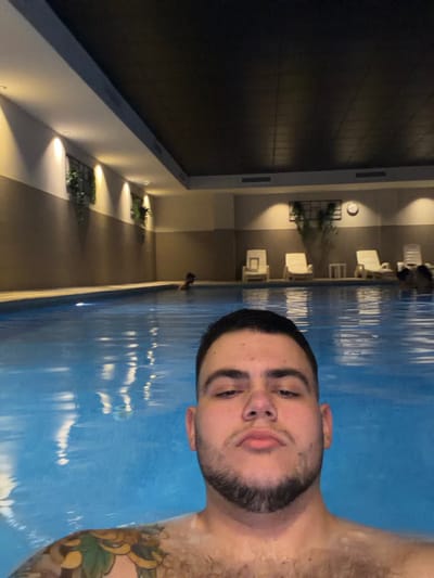 The image displays an indoor swimming pool setting located in Villajoyosa/la Vila Joiosa, Spain. In the foreground, a male individual with short dark hair, a beard, and a visible multi-colored tattoo on his left shoulder is submerged in clear blue water, looking directly forward. In the mid-ground, two other individuals are partially visible within the pool water: one to the left and another further back on the right. The pool area walls are two-toned, featuring a lighter upper section and a darker lower section, illuminated by recessed wall lights. Decorative elements include wall-mounted planters with greenery and a large plant arrangement with a wall clock on the right side. Several white lounge chairs and small side tables are arranged around the perimeter of the pool. The ceiling is dark and textured, with visible air vents along the wall junction.