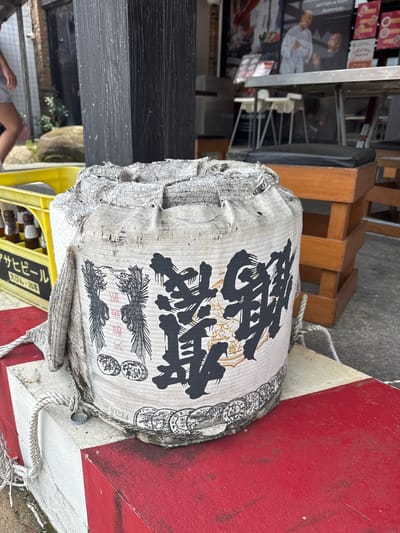 A large, cylindrical object covered in a coarse, white fabric is the central subject of this image. The fabric is frayed and worn, with visible seams and openings at the top. Bold, black Japanese calligraphy adorns the fabric. Two illustrated phoenix-like birds, one on each side of the calligraphy, are also depicted in black. The object is resting on a low, red and white striped structure that appears to be a concrete barrier. A thick, white rope is tied around the object and secured to the barrier. To the left of the central object, a yellow plastic crate containing several glass bottles with brown liquid and white labels is visible. The background shows an outdoor setting with a person walking in the far left, blurred out. Further back, a building with windows and posters, as well as wooden tables and chairs, suggest an establishment like a restaurant or bar. The overall impression is of a rustic, possibly traditional display or storage method for the item.