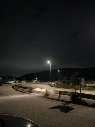 The image presents a night view of a winding road in Anatoliki Attiki, Greece, likely along a coastline or mountainous area. The shot is taken from a low angle, possibly from a vehicle, with the front corner of a car visible in the bottom right.

It is clearly nighttime, with a dark, overcast sky. A prominent, brightly lit streetlight stands in the mid-ground, illuminating a significant section of the asphalt road that curves to the left. Several other streetlights are visible further down the road, casting additional pools of light. The road itself is dark grey, with painted white lines discernible in some illuminated sections.

To the left of the road, dark hills or mountains rise, forming a blurred silhouette against the horizon. In the far distance, a body of water, presumably the sea or a bay, can be seen reflecting distant lights from what appears to be a coastal settlement. Parked cars are visible further down the road on the left, illuminated by the streetlights, and faint vehicle lights suggest ongoing traffic in the distance.

On the right side of the road, a rocky embankment covered with sparse vegetation ascends. A metal guardrail runs parallel to the road. Higher up on the embankment, a large, rectangular sign or billboard is visible, covered in extensive, illegible graffiti, though some faint shapes and outlines can be discerned. Shadows of unseen roadside structures are cast long and starkly onto the road surface in the foreground by the bright artificial light. The overall scene is quiet and still, emphasizing the artificial illumination against the natural darkness of the landscape.