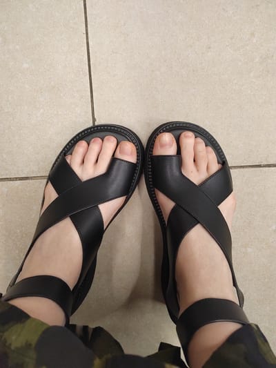 A top-down, medium shot shows a person's feet wearing black cross-strap sandals against a beige tiled floor. The sandals have thick, ridged soles and the straps crisscross over the top of each foot, with one strap encompassing the ankle. The person's toenails are unpolished and their skin is pale. In the foreground, camouflage-patterned fabric is visible, suggesting the lower part of pants or a skirt. The tiled floor appears to be indoors, likely in a residential or public space, with visible grout lines between the tiles. The lighting is even and does not cast strong shadows. There is no indication of the location being in the city of Novogorelovo, Russia.