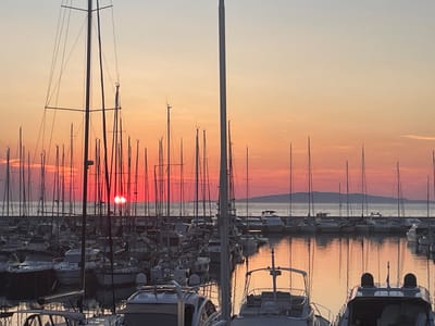 This is a horizontal, eye-level, medium shot of a marina in Punta Ala, Italy, at sunset. The sky is a gradient of pale blue and orange, with the sun visible as a bright, reddish-orange orb partially obscured by boat masts. Numerous sailboats and motorboats are docked in the foreground and middle ground. The water reflects the warm hues of the sunset and the silhouettes of the boats. The scene is serene and peaceful, with no visible people or ongoing activity. The overall impression is one of tranquility and natural beauty.