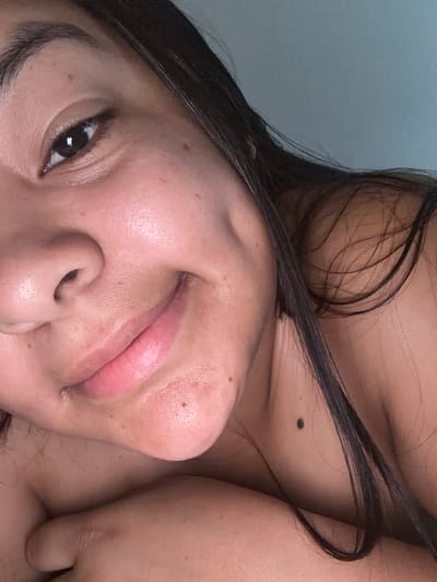 The media file is a close-up, upside-down photograph of a person's face and upper body, with a grey background. The subject has dark hair that is partly draped over their face and shoulder. Their skin appears to have some light freckling and a mole is visible on their neck. The person's eye is dark and open, with visible eyelashes. Their lips are slightly parted and appear to be painted with a light pink lipstick. The overall lighting suggests an indoor setting, possibly with natural light. There are no visible objects or text in the frame. The image captures a moment of quiet introspection or casual pose, with a relaxed and neutral expression. The orientation of the image is unusual, adding an artistic or intimate feel. The location context of Balch Springs, United States of America, does not provide any specific visual cues within the image itself.