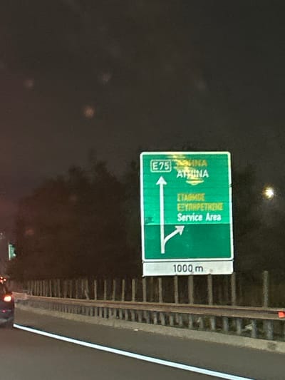 The image captures a highway sign at night, oriented vertically due to the original rotation. The primary subject is a large, reflective green overhead sign, characteristic of European motorways. To its right, a narrower white sign provides additional information. The scene is dark, indicating nighttime, with faint glints of light suggesting reflections from streetlights or passing vehicles, and possibly some lens flare.
The green sign displays directional information in both Greek and English. On the left side, an arrow pointing straight ahead or slightly left indicates "E75" and "ΑΘΗΝΑ ATHINA" (Athens). On the right side, a diverging arrow points towards "ΣΤΑΘΜΟΣ ΕΞΥΠΗΡΕΤΗΣΗΣ Service Area." This suggests a fork in the road where drivers can continue on European Route E75 towards Athens or take an exit for a service area.
The adjacent white sign clearly displays "1000 m," indicating that the upcoming junction or service area is 1000 meters away. A metal guardrail or barrier runs along the right side of the frame, separating the road from the surrounding area. The road surface below the sign is dimly visible. The overall impression is of a driver's perspective approaching a highway exit or service area at night in Agios Stefanos, Greece. No people are visible.