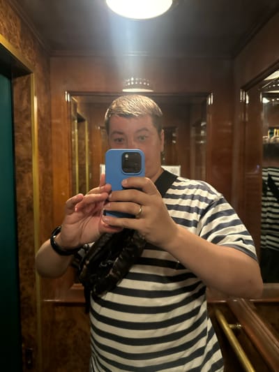 A man is taking a selfie in an elevator with wood-paneled walls. He is wearing a blue and white striped t-shirt and a black fanny pack. He is holding a blue phone in his right hand and wearing a ring on his left ring finger. The elevator appears to be old, with ornate wood paneling and brass accents. The lighting is dim, with a bright light fixture visible on the ceiling. The reflection in the mirror shows another person or object behind the man, also wearing a striped shirt. The location is specified as being in San Martino Buon Albergo, Italy.