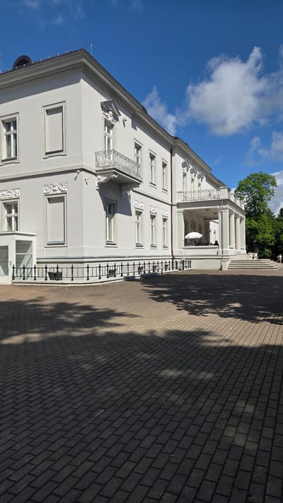 The image depicts a grand, white, neoclassical building with ornate architectural details, including balconies with intricate metal railings and decorative friezes above some windows. The building has multiple floors with evenly spaced rectangular windows. A porch or veranda with white columns and a balcony is visible on the upper level, where a white umbrella and a few people are seated at tables, suggesting an outdoor seating area for a cafe or restaurant.

To the right of the building is a paved walkway made of light brown and dark gray bricks. The walkway is partially covered in dappled shadows cast by trees, indicating it is daytime with a bright sun. The sky is a vibrant blue with scattered white clouds. Lush green trees are visible in the background, behind the building and to the left. A few people are walking up a set of stairs leading to the building's entrance on the far right.

The scene appears to be a sunny day, likely in the late morning or early afternoon, given the strong shadows. The overall impression is one of a serene and elegant public space, possibly a park, a museum, or a historic estate in Palanga, Lithuania. There is no visible text in the image.