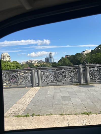 The image captures a view from a car window looking out at a city landscape. In the foreground, a grey stone sidewalk with a decorative inlaid brick path runs horizontally. Behind the sidewalk is an ornate grey metal railing with intricate scrollwork and circular emblems. Beyond the railing, a body of water, likely a river, is visible. The background features a cityscape with lush green trees, several modern buildings of varying heights, including a tall, slender skyscraper and a lighter-colored, stepped building. The sky is a bright blue with scattered white clouds, indicating a clear day. The overall scene suggests a well-maintained urban area with a mix of architectural styles, possibly near a riverfront promenade in Tbilisi, Georgia.
