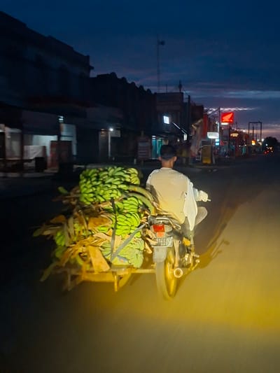 An elevated rear-view photograph captures a male individual operating a motorcycle with an attached sidecar on an asphalt road at twilight or dawn in Grong-Grong, Indonesia. The rider, observed from behind, is wearing a light-colored long-sleeved shirt. The motorcycle displays a license plate reading '5141 NM 08.22'. The sidecar is heavily laden with numerous bunches of unripe green bananas and dried plant material, secured by visible restraints. The immediate foreground of the road is brightly illuminated by a strong yellow light source, casting elongated shadows of the vehicle and rider. The sky above is dark blue, featuring scattered clouds with subtle reddish-purple twilight hues. Structures consistent with commercial buildings or residences line both sides of the street. On the right side of the road, multiple illuminated signs are visible, including a prominent red Honda logo, a 'BSI ATM' sign, and a sign identifying 'LAXANA MOTOR'. Streetlights are active along the thoroughfare in the distance.