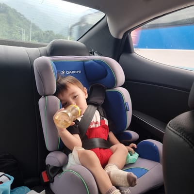 A toddler is seated in a DAIICHI brand car seat, drinking from a yellow bottle. The toddler is wearing a red garment over a white t-shirt and beige socks. The car seat is gray, blue, and green, and secured with black straps. The scene is inside a vehicle, with a view of a green, mountainous landscape through the rear window. The location is Ucheon-myeon, South Korea.