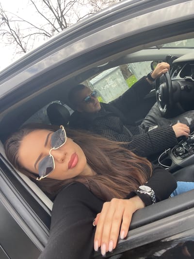 This image shows two people inside a car. The car appears to be parked, based on the tree branches visible through the open roof. The individual closest to the camera is a woman wearing sunglasses, a black top, and a watch on her left wrist. Her hand rests on the car's interior. Behind her, partially visible, is a man also wearing sunglasses and a black jacket. He is holding the steering wheel. The background suggests an outdoor setting, possibly in a residential area. There is no evidence to suggest that the image was taken in Pchelishte, Bulgaria.