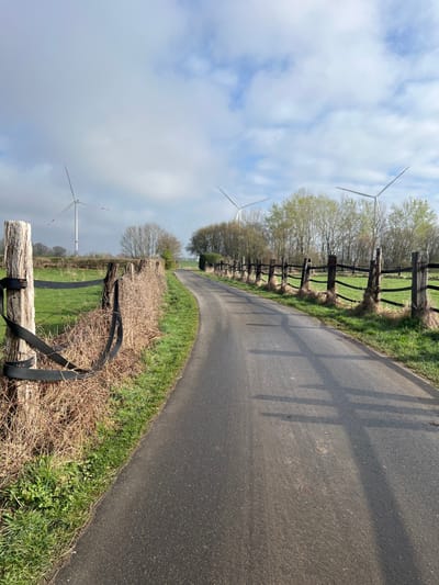 The image depicts a rural scene in Würselen, Germany, featuring an asphalt road curving gently towards the horizon under a partly cloudy sky. The road, dark gray in color, is free of vehicles or pedestrians. On the left, a weathered wooden fence post is visible in the foreground, with a black strap loosely wrapped around it. Behind this post, a dense, dry hedge, possibly a hedgerow, runs parallel to the road, separating it from a vibrant green grassy field. Further back on the left, a large wind turbine with white blades and a red tip on one blade stands prominently against the sky.
On the right side of the road, a fence constructed from dark wooden posts and horizontal rails defines the edge of another green grassy field. Shadows from these fence posts are cast diagonally across the asphalt road towards the left. Beyond the fences and fields, deciduous trees with nascent green foliage indicate early spring conditions. Two additional wind turbines are visible in the mid-distance; one on the right, partially obscured by trees, and another on the left, further down the road's path, both exhibiting white blades. The sky above is a mix of bright blue and scattered white cumulus clouds. No individuals are present in the scene.