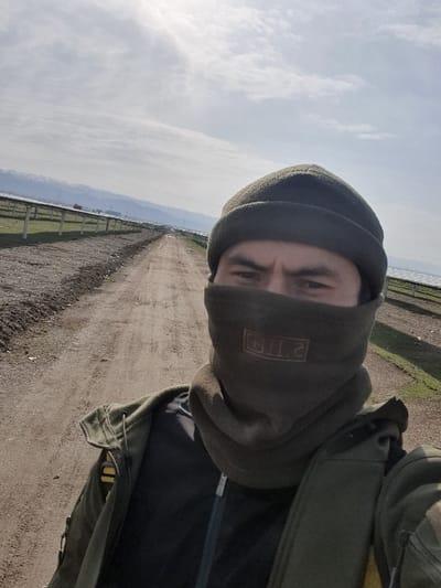 A person wearing a dark beanie and a face covering with the text "5.11" stands on a dirt road. The person is wearing a dark jacket. The road stretches into the distance, flanked by a fence on one side and fields on the other. The sky is partly cloudy.