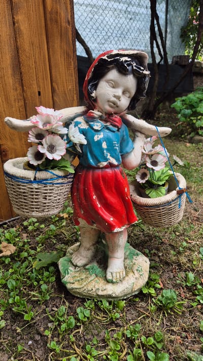 A weathered garden statue of a young girl carrying baskets of artificial flowers on her shoulders stands on a patch of green ground. The statue is made of concrete and is painted with red, blue, and white colors. The girl wears a red hood, a blue shirt, and a red skirt. Her face is tilted down, and her eyes are closed. The baskets she carries are woven from natural materials and contain white artificial flowers with pink tips and green leaves. The statue is placed in a garden setting, with a wooden fence to the left and trees and foliage in the background. The ground is covered in grass and soil, with some fallen leaves. The overall scene suggests a rustic and somewhat neglected outdoor space, possibly in a residential area. The image is taken at a medium distance, with the statue being the main focal point. No specific location in Doirentsi, Bulgaria is identifiable from the image itself.