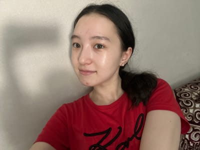 A young woman with dark hair pulled back in a ponytail and wearing a red t-shirt is looking towards the camera. She has light-colored skin with some minor blemishes and is wearing a small pearl earring. The background appears to be a wall with textured wallpaper and a shadow cast on it. Part of a patterned cushion is visible on the right side of the frame. The overall scene suggests a casual indoor setting. The image does not provide any specific details indicating a location in Nookat, Kyrgyzstan.