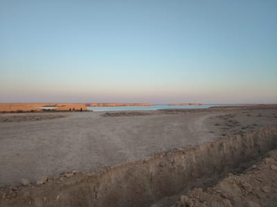 In the foreground, a trench is visible, its edges eroded and littered with rocks and soil. The ground surrounding the trench is dry and cracked, with sparse vegetation. In the mid-ground, a group of three individuals are standing on a sandy plain. Beyond them, a body of calm water, possibly a lake or sea, stretches towards the horizon. The distant shore is characterized by low, arid cliffs. The sky above is clear and exhibits a gradient of soft blues and pale pinks, indicative of either sunrise or sunset. The overall environment appears to be a desert landscape situated in or near Haratan, Uzbekistan.