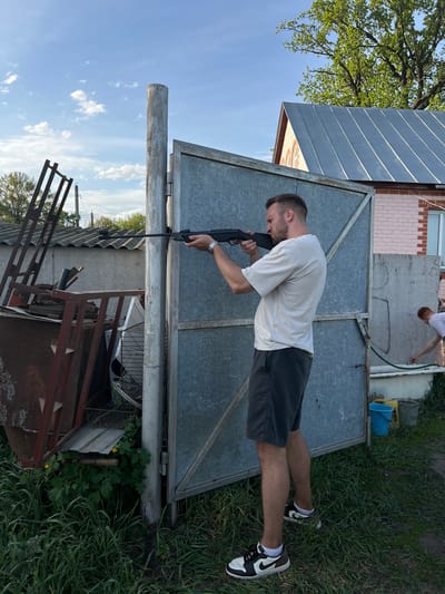 The photograph depicts a man holding a rifle outdoors. He wears a white t-shirt, dark shorts, and black and white shoes. The man is positioned in front of a large metal gate, with a small brick building visible in the background. The surrounding environment includes grass, foliage, and miscellaneous metal structures. The location is Слобода, Russia.