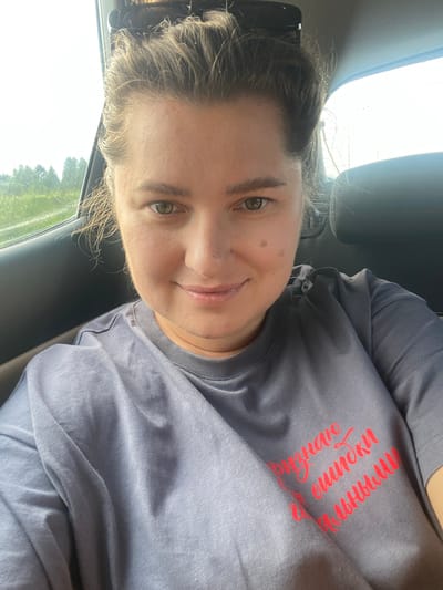 A woman with light brown hair pulled back is in the backseat of a car, looking at the camera. She is wearing a grey t-shirt with red lettering that reads "Признаю ошибки полными" which translates to "I admit my mistakes fully." The woman has green eyes and a light complexion. The background outside the car window shows a green, grassy field with trees in the distance, suggesting a rural or suburban setting near Залесная, Russia. The interior of the car is visible, with a dark seat behind her and a window to her left. The overall lighting suggests it is daytime, likely with overcast or bright skies.