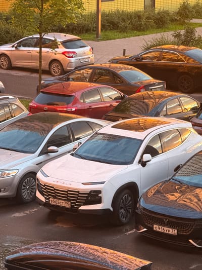 This image depicts a parking lot in Drozhzhino, Russia, bathed in the warm glow of sunset. Several cars are parked, including a silver hatchback with a license plate starting with "P416HK", a red hatchback with "KIA" badging, a dark sedan reflecting the sunset, and a white SUV with a license plate "E565CT 790". In the foreground, a dark roof box is visible. The scene is surrounded by green foliage and a building with a yellow, textured facade. The lighting suggests it is late afternoon or early evening, casting long shadows and highlighting the metallic surfaces of the vehicles.