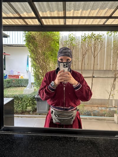 The image captures a mirror selfie of an individual in Ban Huai Phlap, Thailand. The person is seen from the upper thigh upwards, wearing a camouflage-patterned balaclava that conceals the head and face except for the eyes, which are made up with dark eyeliner. The individual wears a maroon bomber-style jacket with white and dark blue striped cuffs and collar over a dark garment, and a gray fanny pack with white zippers is secured around the waist. A silver-colored wristwatch is visible on the left wrist. Both hands hold a light-colored smartphone with a clear case, displaying "251" text on its side, pointed towards the mirror to capture the reflection.

The background, reflected in the mirror, reveals an outdoor setting beneath a translucent corrugated roof supported by dark metal beams. To the left, a manicured lawn with hedges and a light-colored structure is visible, alongside a partial view of a white building with a blue element. Centered behind the individual is a lush, mature green tree. To the right, several young, sparse trees stand in front of a corrugated metal wall. The ground in the reflected scene appears to be concrete or paved. The actual foreground of the image consists of a dark, granular surface, likely the mirror's frame or a counter.