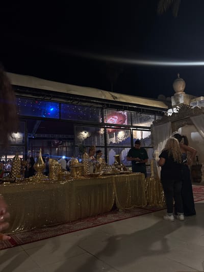The image shows an outdoor event at night at a restaurant in Ramla, Israel, located at 31.926 latitude and 34.873 longitude. There are three people standing near a dessert buffet table covered with a gold sequined tablecloth. The buffet is decorated with several gold vases and serving platters. The background shows a glass-walled structure, partially obscured by a white canopy. There are a few people visible inside, but they are out of focus. Behind the canopy is a wall with a traditional architectural element resembling a minaret or a dome. The environment is dimly lit and illuminated by spotlights. The ground is paved with square tiles. The mood is festive, celebratory, and relaxed.