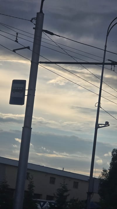 The image depicts an outdoor urban environment in Nizhnyaya Maktama, Russia, captured from a low-angle perspective looking upwards towards the sky. Two prominent utility poles dominate the foreground. The pole on the left is a grey metallic structure, featuring multiple black overhead power or communication lines crisscrossing the frame. Attached to this pole is a rectangular blue and white sign, indicating a bus stop, with a stylized bus silhouette. The pole on the right is a darker, slenderer utility pole, possibly a street light pole, also supporting several overhead wires. A surveillance camera is mounted on a horizontal bracket extending from this right pole, aimed towards the left.
The sky occupies the majority of the frame, displaying a transition of cloud types: dense, dark grey clouds are visible in the upper portion, gradually lightening to a mix of white and pale yellow clouds closer to the horizon line, suggesting either dawn or dusk. Several small dark specks, likely birds, are faintly visible against the sky. Below the cloud line and partially obscured by the utility poles and foreground elements, a light-colored, multi-story building with a flat roof and multiple rectangular windows is visible in the midground. Several dark green, coniferous-like trees and bushes are situated in front of the building. A faint, partial "VA" logo is visible on a sign or structure near the base of the left utility pole. No people are present in the scene.