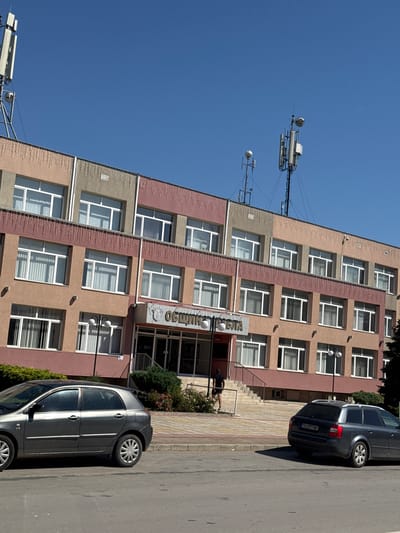 In Shabla, Bulgaria, a three-story municipal building with a light brown, pink, and grey facade stands under a clear blue sky. The building features multiple windows with white frames on each level, and some windows have vertical blinds. A sign above the entrance reads "ОБЩИНА ШАБЛА" (Municipality of Shabla). A person wearing a black t-shirt and shorts is walking up a set of stairs to the building's entrance. Two cars are parked on the street in front of the building; a dark grey hatchback is on the left, and a dark grey station wagon is on the right, displaying a license plate with "TX 2977 MT". Several communication towers with antennas are visible on the roof of the building and in the background. Green bushes and some flowering plants are situated in front of the building.
