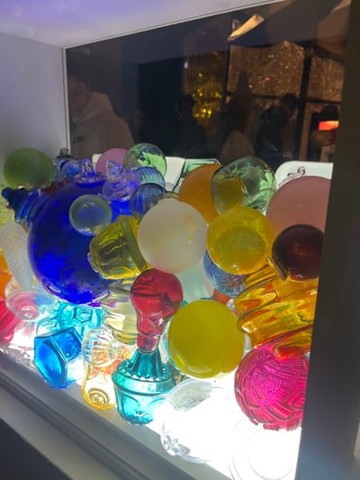 This image captures an illuminated display of numerous multi-colored glass objects, predominantly spheres and abstract shapes, tightly packed within a confined space. The objects, ranging in color from deep blue, light blue, green, yellow, orange, red, pink, white (frosted), and clear, are lit from below, causing them to glow. A significant portion of the scene consists of reflections on the upper glass or transparent surface of the display.

Reflected in the upper left, a person, likely an adult, is visible from the chest up, wearing a light-colored hooded sweatshirt. This individual appears to be looking towards the display. Further reflections in the background show indistinct silhouettes of other individuals and blurred yellow-orange lights, possibly originating from interior fixtures or a street scene beyond the immediate display. The overall environment, identified as a location in the city of Sélestat, France, appears to be an indoor or semi-enclosed public space, possibly a shop window or art installation, observed at night or in low light conditions. No direct interactions between individuals are discernible; the primary interaction captured is the observation of the display.