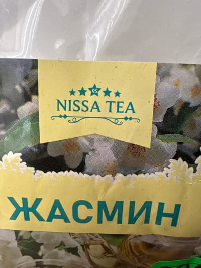 The image is a close-up of a yellow label on a product, likely tea packaging, featuring text in both English and Cyrillic script. The English text, appearing in a teal color, reads "NISSA TEA" and is accompanied by three teal stars and a decorative flourish. Below this, in larger teal Cyrillic letters, the word "ЖАСМИН" is prominently displayed. The background of the label features an image of white flowers, possibly jasmine, interspersed with green leaves. The label itself is slightly torn along the bottom edge and has a scalloped border. The overall image quality suggests it was taken with a mobile phone camera. There is no visible indication of the city of Izyayu, Russia, or any specific environmental context beyond the product packaging.
