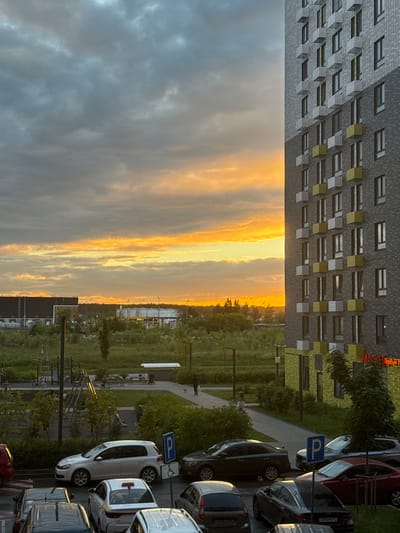 This is a vertical photograph of a residential building and a parking lot at sunset, likely taken in Drozhzhino, Russia. The building is modern, with a facade of gray brick and a repeating pattern of windows and balconies, some of which are accented with yellow. To the right of the building is a parking lot filled with various cars. One car is white, and another is dark colored, with a parking sign displaying a blue 'P' symbol visible above them. A small, pale-colored figure, possibly a person, can be seen walking on a paved path in the middle ground, near some green foliage. The sky is dramatic, with a striking contrast between the dark, brooding clouds on the left and the vibrant, fiery orange and yellow hues of the sunset on the right, indicating the time of day. The overall mood is tranquil, with the setting sun casting a warm glow over the scene. No specific event appears to be taking place, suggesting a typical evening in this suburban or semi-urban setting.