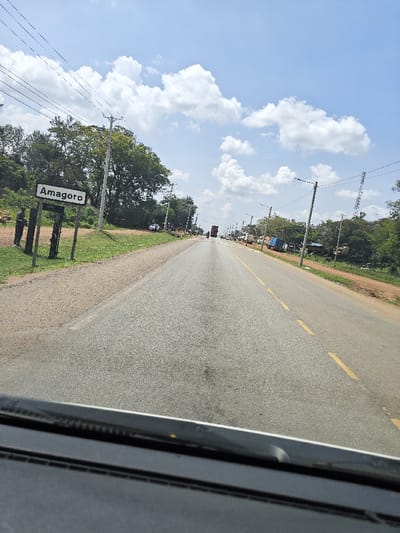 A road stretches ahead under a partly cloudy sky, marked with intermittent yellow lines. A road sign indicating "Amagoro" stands to the left, near figures standing on the roadside. Utility poles line the route, and vehicles are visible further along the road, suggesting a populated area. The view is from within a vehicle, focusing on the road ahead.