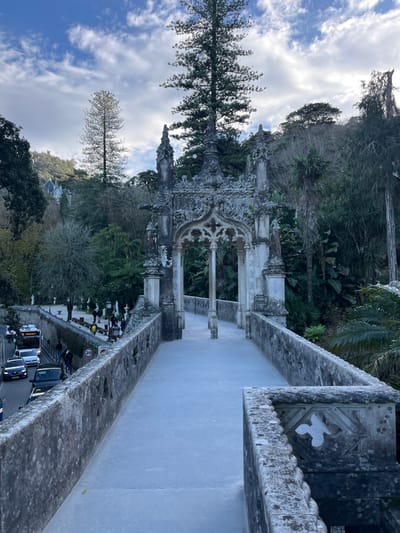 A historic, ornate stone structure with arches and columns stands amidst lush greenery, framed by a bright sky with scattered clouds. The structure overlooks a road with parked vehicles. A raised walkway with detailed stone railings runs parallel. The architecture is aged, possibly from a castle or palace. Tall trees punctuate the landscape, contributing to a vibrant and diverse natural backdrop.