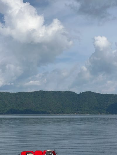 The image displays a wide, calm body of water, likely a lake, under an overcast sky in the City of Unknown, El Salvador. The upper approximately 40% of the frame is filled with a cloudy sky, featuring a mix of bright white, puffy clouds and diffused grey clouds, with hints of pale blue sky visible between them. Below the sky, a continuous expanse of densely forested, dark green mountains or hills stretches across the midground, forming a distant horizon. Small, indistinct structures are faintly visible along the upper ridgeline of these mountains.

Beneath the mountains, the wide body of water exhibits a grey-blue hue, reflecting the sky, and is characterized by numerous uniform, parallel horizontal ripples across its surface, suggesting calm conditions. A narrow, lighter-toned strip of distant shoreline with scattered small buildings is discernible between the water and the base of the mountains.

In the immediate foreground, occupying the bottom portion of the image, are the rear sections of two bright red watercraft, appearing to be a catamaran-style pedal boat or a similar small recreational vessel. The left red section shows a beige or light-colored seat. The right red section features a black steering wheel positioned at a white or light-colored console, also with a beige seat visible. These watercraft are stationary on the water, with no individuals visible on board or in the immediate vicinity.